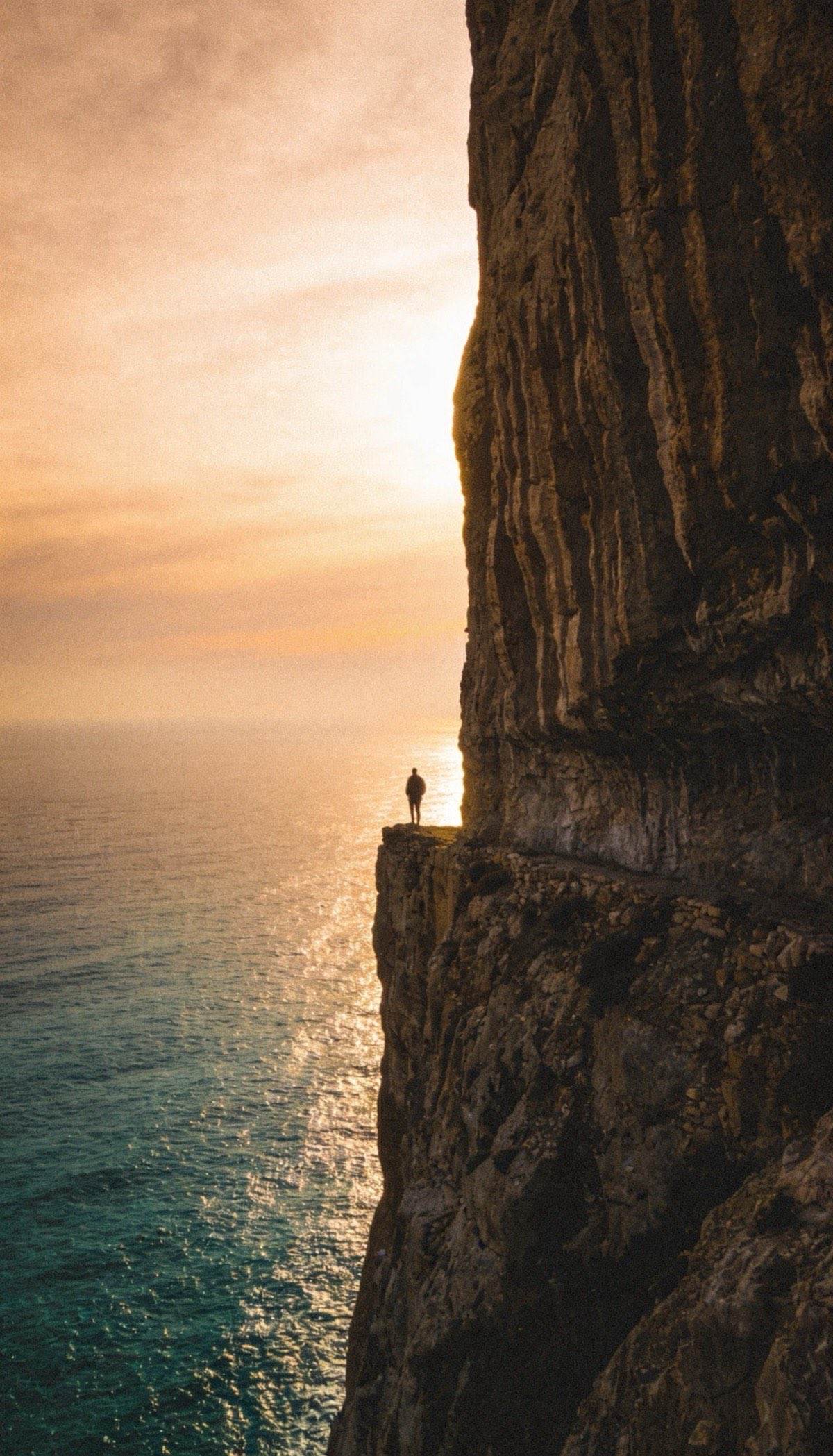 Pessoa sozinha na beira de uma falésia olhando o mar ao pôr do sol